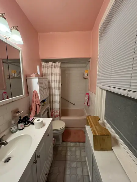 Bathroom before the remodel: pink walls, a pink tub with a striped shower curtain, small white wall tile, and vinyl floor tile.