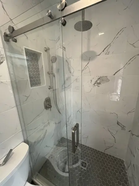 Finished shower with white marble-look wall tile, gray hexagon mosaic floor and niche, a corner bench, rain showerhead, handheld sprayer, and a frameless sliding glass door.